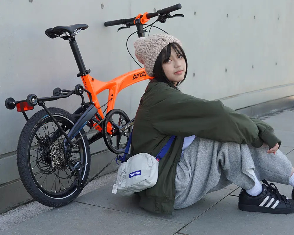 Neon Orange Birdy URB 9-speed folding bike parked against a concrete wall, showing the standard trolley rack with rolling wheels