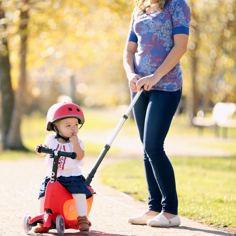 Toddler sitting on a red Micro Mini2go while a parent pushes with the push bar in the park