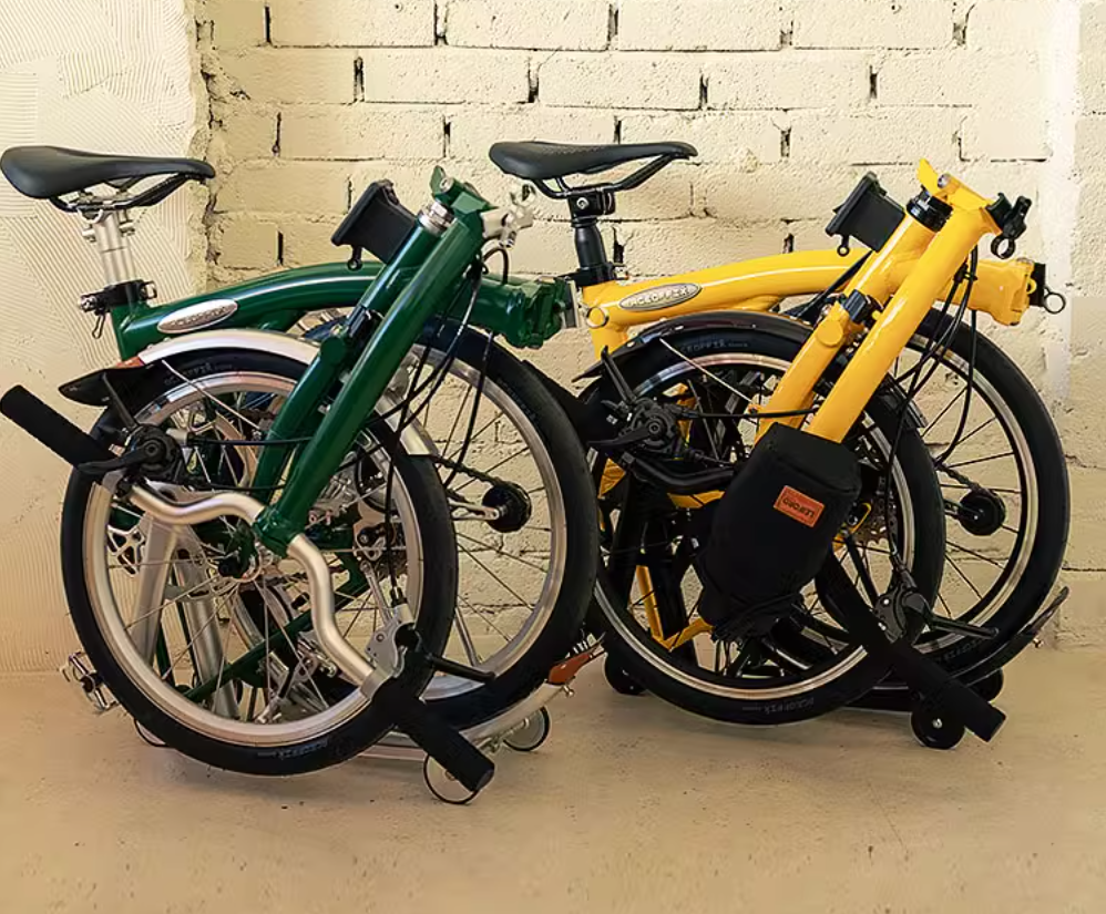 Two Aceoffix Y5 tri-fold bikes in green and yellow, folded side by side against a brick wall