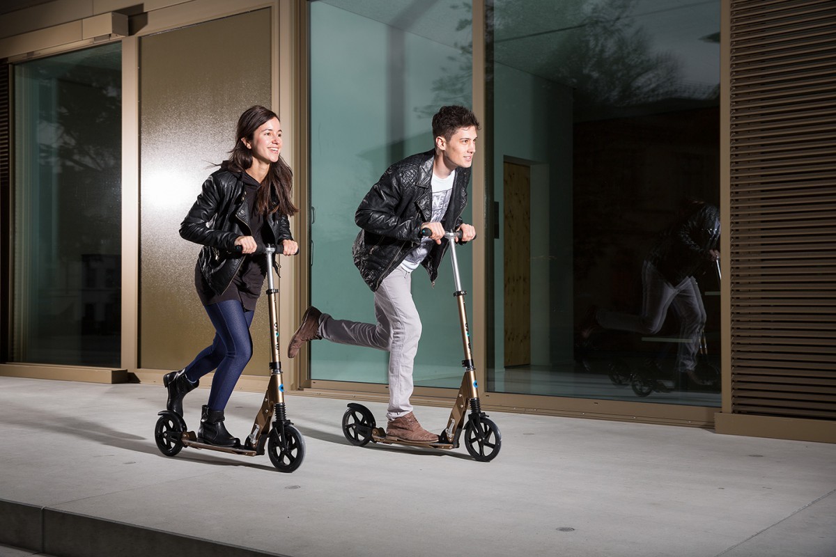 Two young riders on copper Micro Suspension 200mm scooters gliding along a pavement outside a building
